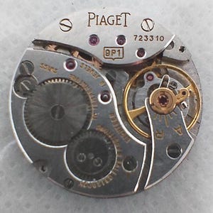 May include: Close-up of a watch movement with the brand name "Piaget" in silver lettering. The movement features intricate gears, jewels, and the text "Swiss". The model number "723310" is visible.