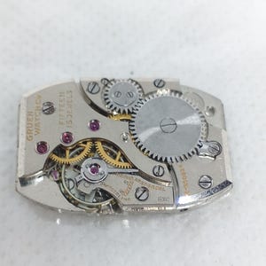 May include: Close-up of a Gruen Watch Co. watch movement with visible gears, jewels, and text. The movement is rectangular, with the words "Fifteen 15 Jewels" and "Swiss" visible. The watch mechanism is silver-toned.