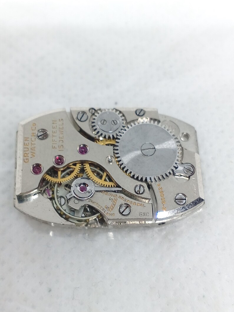 May include: Close-up of a Gruen Watch Co. watch movement with visible gears, jewels, and text. The movement is rectangular, with the words "Fifteen 15 Jewels" and "Swiss" visible. The watch mechanism is silver-toned.