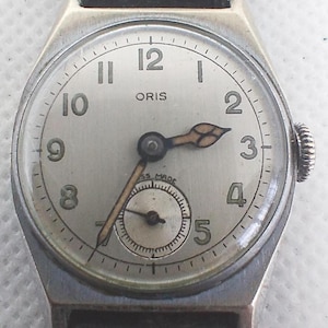 Peut inclure: Une montre-bracelet Oris vintage avec un cadran argenté, un bracelet en cuir noir et un petit cadran des secondes. La montre présente des chiffres noirs, des aiguilles dorées et une couronne. Le cadran mesure environ 3,2 cm de diamètre.