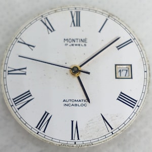 Może przedstawiać: Zegarek w stylu vintage z białym tłem, czarnymi rzymskimi cyframi i czarnymi wskaz&oacute;wkami. Tarcza zegarka zawiera napis "MONTINE 17 JEWELS AUTOMATIC INCABLOC" oraz okienko daty z numerem 17.