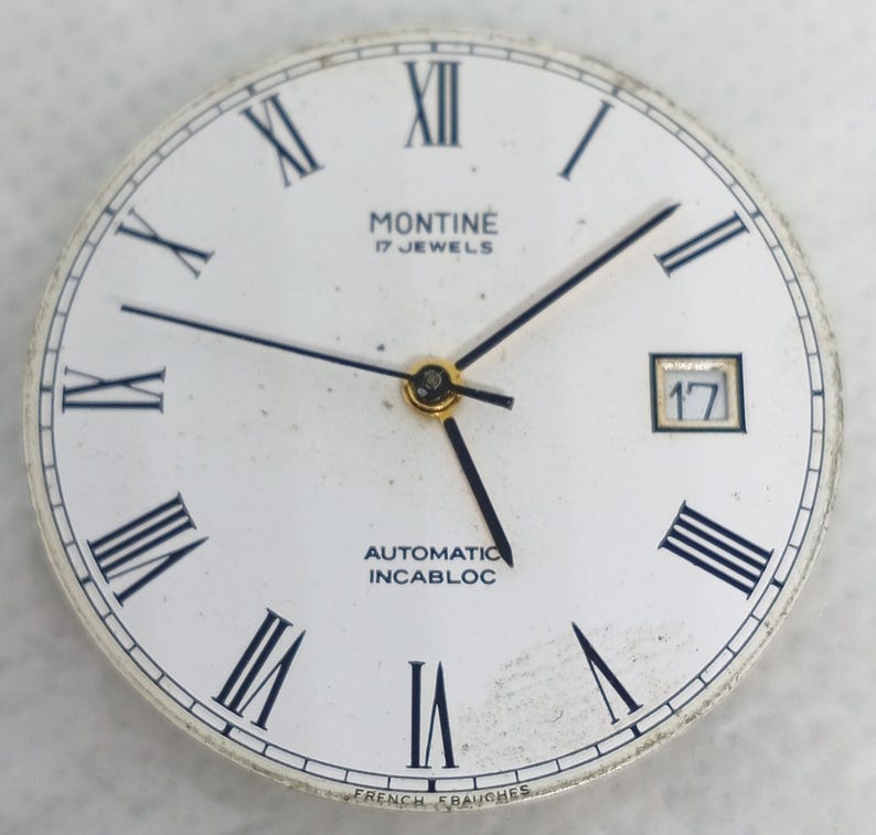 Może przedstawiać: Zegarek w stylu vintage z białym tłem, czarnymi rzymskimi cyframi i czarnymi wskaz&oacute;wkami. Tarcza zegarka zawiera napis "MONTINE 17 JEWELS AUTOMATIC INCABLOC" oraz okienko daty z numerem 17.
