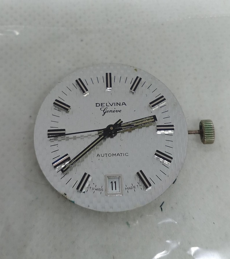 May include: A silver watch face with black and silver hour markers and hands. The watch face features the text "DELVINA Gen&egrave;ve AUTOMATIC" and a date window showing the number 11. A textured crown is visible on the right side.