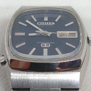 May include: A vintage Citizen Electronic Cosmotron watch with a rectangular silver case and a dark blue face. The watch face displays the day and date, with the brand name and model details visible. The watch has silver hands and markers.