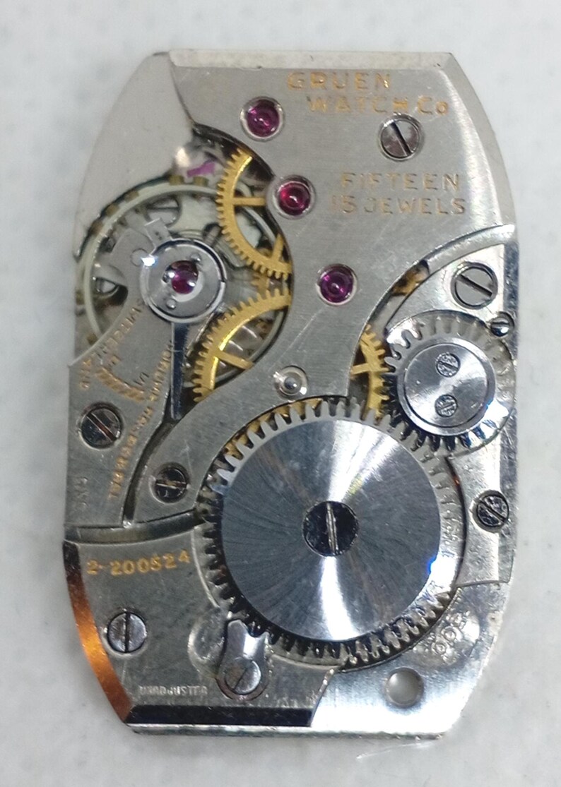 May include: Close-up of a Gruen Watch Co. watch movement. The silver-toned mechanism features gears, jewels, and screws. The text "FIFTEEN 15 JEWELS" is visible. The watch movement is rectangular with rounded edges.