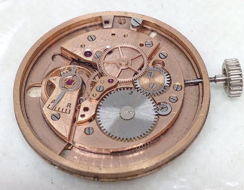 May include: Close-up of a watch movement with intricate gears, screws, and a winding stem. The metal components are in shades of gold and silver, with small red jewels. The watch mechanism is round and detailed.
