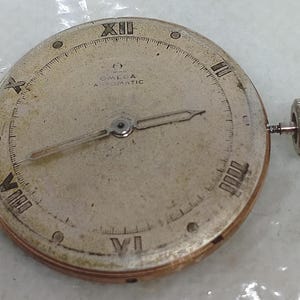 May include: A vintage Omega Automatic watch face with Roman numeral hour markers and delicate hands. The silver-toned face shows signs of age, with the Omega logo and text visible. A silver crown is attached to the side.