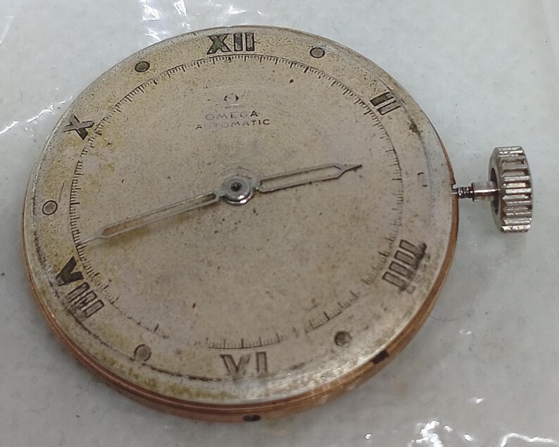May include: A vintage Omega Automatic watch face with Roman numeral hour markers and delicate hands. The silver-toned face shows signs of age, with the Omega logo and text visible. A silver crown is attached to the side.