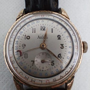 Op de afbeelding: Een vintage goudkleurig Amirol polshorloge met een zilveren wijzerplaat, zwarte leren band en een datumweergave. Het horloge heeft een subwijzerplaat en een draaiende buitenring met dagmarkeringen. De wijzers zijn goudkleurig.