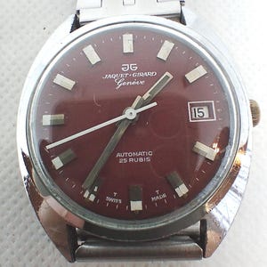 May include: A vintage Jaquet Girard Genève wristwatch with a burgundy face and silver hands. The watch features a date display at the 3 o'clock position and the text "AUTOMATIC 25 RUBIS". The watch has a silver metal strap.