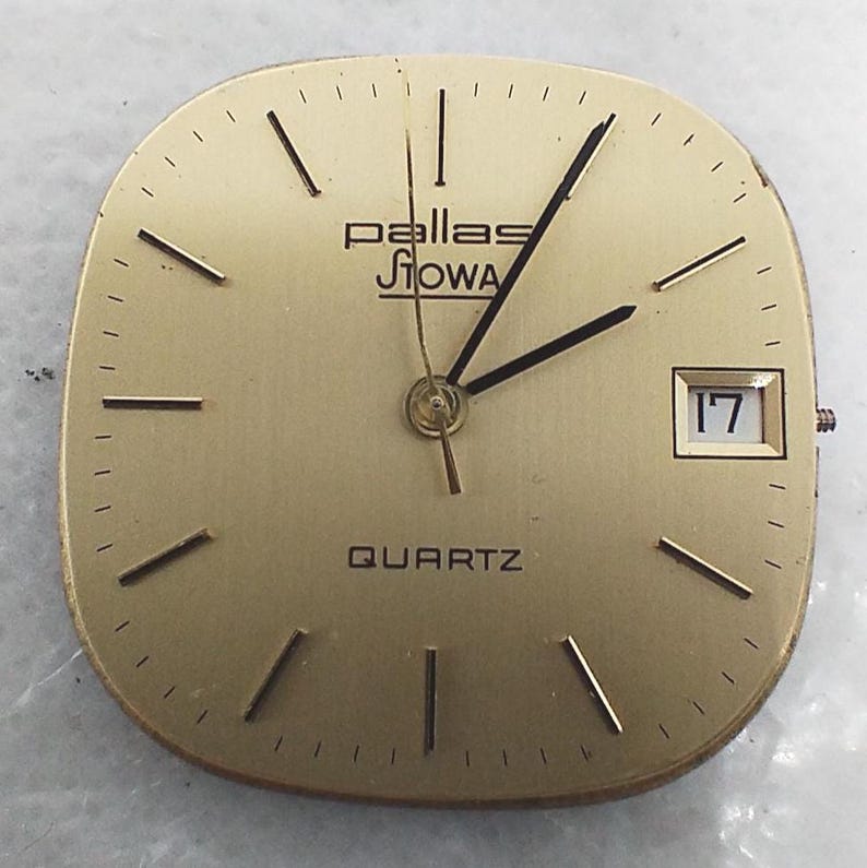 May include: A gold-toned watch face with the brand name "Pallas Siowa" and the word "QUARTZ" at the bottom. The watch face has black hands and markers, with a date window displaying the number "17".
