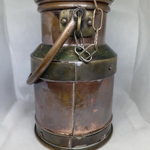 Op de afbeelding: Een vintage melkkan van koperkleurig metaal met een scharnierend deksel en een handvat. De kan heeft een ketting- en sluiting, en een groene band aan de bovenkant. Het metaal heeft een verweerde uitstraling.