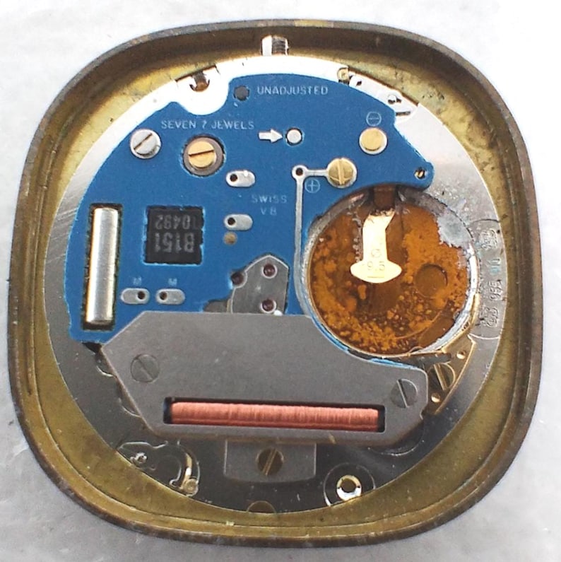 May include: Close-up of a watch movement with a blue circuit board and gold-colored metal casing. The board has the text "SEVEN 7 JEWELS" and "SWISS V8". A battery and other small components are visible.