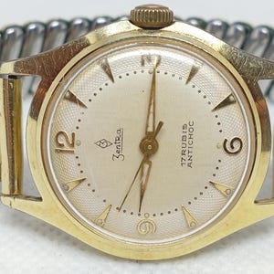 Op de afbeelding: Een vintage goudkleurig polshorloge met een crèmekleurige wijzerplaat. Het horloge heeft een gouden lunette, wijzers en uurmarkeringen. De horlogeband is een combinatie van goud- en zilverkleurig metaal. De wijzerplaat leest "Zentra" en "17 Rubis Antichoc".