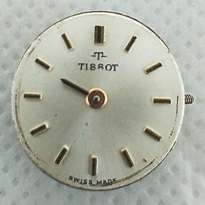 Op de afbeelding: Een zilverkleurige Tissot wijzerplaat met goudkleurige wijzers en uurmarkeringen. De wijzerplaat heeft de merknaam "TISSOT" in zwart, en "SWISS MADE" staat onderaan. Een kleine schroef is zichtbaar aan de zijkant.