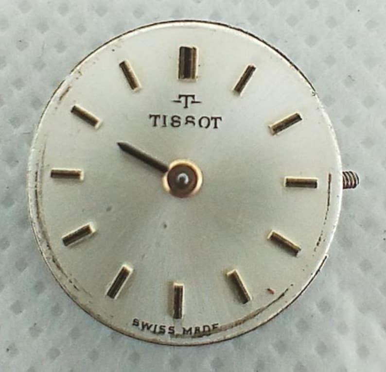 Op de afbeelding: Een zilverkleurige Tissot wijzerplaat met goudkleurige wijzers en uurmarkeringen. De wijzerplaat heeft de merknaam "TISSOT" in zwart, en "SWISS MADE" staat onderaan. Een kleine schroef is zichtbaar aan de zijkant.