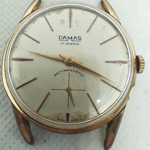 Può includere: Un orologio da polso DAMAS vintage color oro con quadrante color crema. L'orologio presenta lancette e indici dorati, il testo "DAMAS", "17 JEWELS" e "ANTIMAGNETIC". L'orologio ha un quadrante rotondo e una cassa color oro.