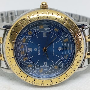 Op de afbeelding: Een Junghans Astro-Chron polshorloge met een blauwe wijzerplaat voorzien van dierenriemsymbolen, Romeinse cijfers en een datumvenster. Het horloge heeft een goudkleurige ring met 24-uurs aanduiding en een tweekleurige metalen schakelband.