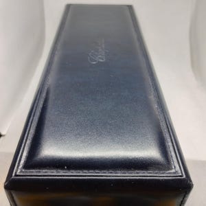 Może przedstawiać: Prostokątne, granatowe pudełko z przeszytą krawędzią. Pudełko ma błyszczące wykończenie i srebrny napis "Chopard" na górze. Pudełko jest prawdopodobnie przeznaczone na biżuterię lub zegarek.