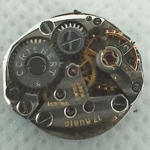 Op de afbeelding: Close-up van een uurwerk met zichtbare tandwielen, schroeven en de merknaam "CORTEBERT" op een cirkelvormig onderdeel. Het uurwerk heeft een complexe opstelling van metalen onderdelen, waaronder goudkleurige tandwielen en kleine rode juwelen. De tekst "17 RUBIS" is ook zichtbaar.