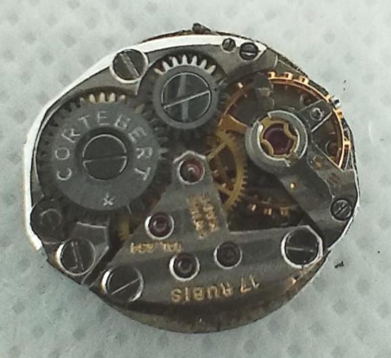 Op de afbeelding: Close-up van een uurwerk met zichtbare tandwielen, schroeven en de merknaam "CORTEBERT" op een cirkelvormig onderdeel. Het uurwerk heeft een complexe opstelling van metalen onderdelen, waaronder goudkleurige tandwielen en kleine rode juwelen. De tekst "17 RUBIS" is ook zichtbaar.
