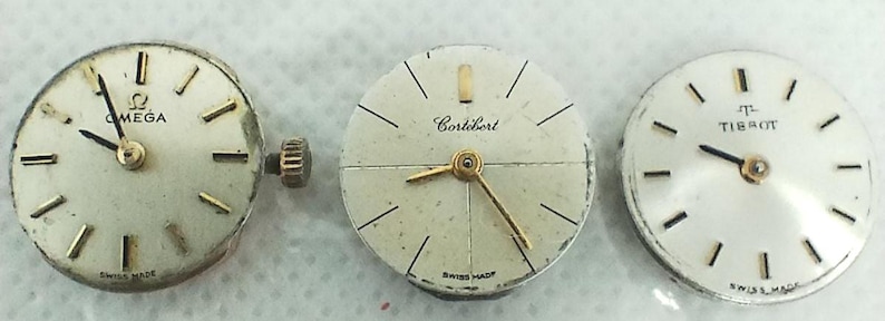 Op de afbeelding: Drie vintage horlogezichten: Omega, Cort&eacute;bert en Tissot. Elk horlogezicht heeft een witte achtergrond met goudkleurige wijzers en markeringen. Het Omega-horlogezicht heeft een gouden kroon. Alle drie zijn gemarkeerd met "Swiss Made".