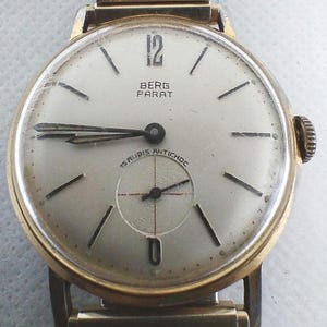 Peut inclure: Une montre-bracelet dorée avec un cadran blanc et des aiguilles noires. Le cadran de la montre porte le texte "BERG PARAT" et "15 RUBIS ANTICHOC".