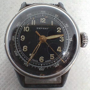 Könnte beinhalten: Eine Vintage-Armbanduhr mit schwarzem Zifferblatt und goldenen Zeigern. Das Zifferblatt hat weiße Ziffern und das Wort "EXPERT" über der Mitte. Die Uhr hat eine silberne Lünette und ein schwarzes Lederarmband. Die Uhr ist mit "SWISS MADE" gekennzeichnet.