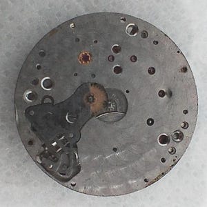 May include: Close-up of a vintage watch movement. The circular, silver-toned mechanism features numerous small holes, gears, and intricate components. A dark, irregularly shaped piece is attached to the main body, with a small, reddish-brown jewel visible.