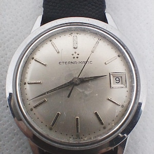 Puede incluir: Un reloj de pulsera Eterna-Matic plateado con una correa de cuero negro. La esfera del reloj es plateada con manecillas e indicadores de hora negros. La fecha se muestra en la posición de las 3 en punto. El reloj tiene el texto "ETERNA-MATIC" en la esfera.