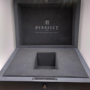 May include: Open grey watch box with the Perrelet logo and text. The box is lined with grey fabric and has a square compartment. The text reads "PERRELET 1777 INVENTOR OF THE AUTOMATIC WATCH."