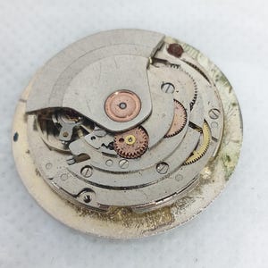 Może przedstawiać: Zbliżenie mechanizmu starego zegarka, ukazujące wewnętrzne mechanizmy. Okrągły mechanizm prezentuje skomplikowany układ k&oacute;ł zębatych, sprężyn i metalowych element&oacute;w w odcieniach srebra, złota i miedzi. Szczeg&oacute;łowy widok inżynierii zegarmistrzowskiej.
