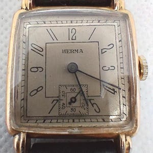 Könnte beinhalten: Eine vintage goldfarbene Armbanduhr mit rechteckigem Zifferblatt und schwarzem Lederarmband. Das Zifferblatt zeigt den Markennamen "HERMA", schwarze Ziffern und ein kleines Sekunden-Zifferblatt. Die Uhr hat eine goldene Krone und ein klassisches Design.