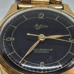 May include: A gold-tone wristwatch with a black circular dial featuring the text Bifora, HOCKPROOF, and 16 RUBIS. The watch face displays gold-colored hour markers, numerals at 12, 3, 6, and 9, and thin gold hands, all encased in a gold-tone metal frame.