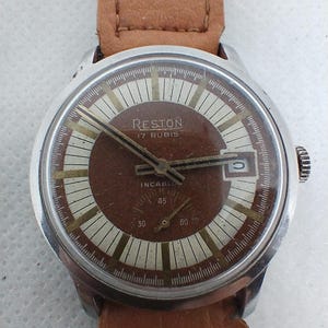 Peut inclure: Une montre-bracelet argentée avec un cadran brun et un bracelet en cuir brun. Le cadran de la montre a des marques blanches et le texte "RESTON 17 RUBIS INCABLOC 45" imprimé dessus.