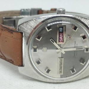 Op de afbeelding: Een zilverkleurig polshorloge met een bruine leren band. De wijzerplaat is grijs met zilveren wijzers en markeringen. De datumweergave toont "SAM 19". Het horloge heeft een getextureerde lunette en een bruine leren band met krokodillenpatroon.