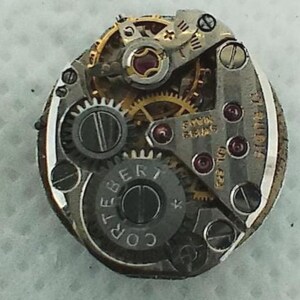Op de afbeelding: Close-up van een uurwerk met ingewikkelde tandwielen, schroeven en juwelen. Het uurwerkmechanisme toont de tekst "SWISS MADE" en "CORTEBERT". De metalen onderdelen zijn zilver- en goudkleurig.