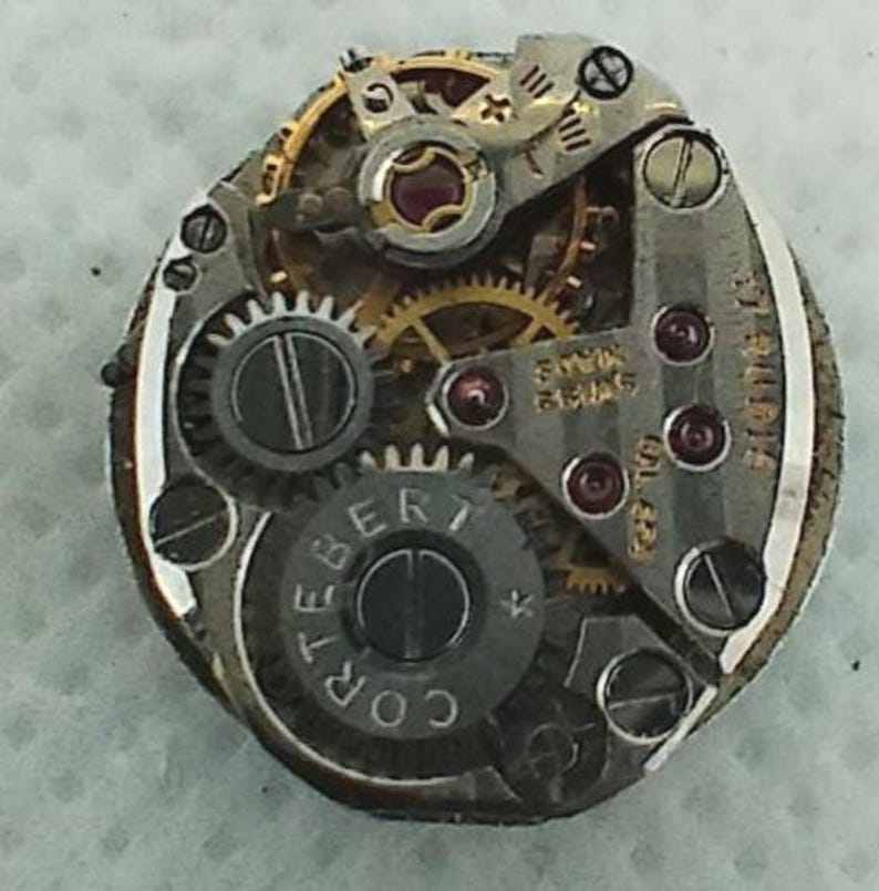Op de afbeelding: Close-up van een uurwerk met ingewikkelde tandwielen, schroeven en juwelen. Het uurwerkmechanisme toont de tekst "SWISS MADE" en "CORTEBERT". De metalen onderdelen zijn zilver- en goudkleurig.