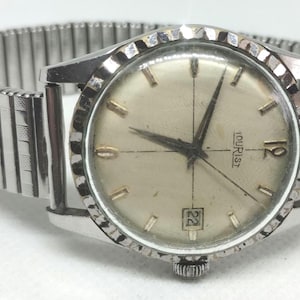 Puede incluir: Reloj de pulsera plateado vintage con esfera clara y visualización de la fecha. El reloj tiene una correa extensible plateada. La esfera del reloj tiene marcadores de hora y agujas doradas. La palabra "TOURIST" está impresa en la esfera.