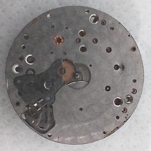 May include: A circular, metallic watch movement component with numerous small holes and embedded jewels. The component features gears, levers, and other mechanical parts. The metal has a worn, aged appearance, suggesting it is vintage or antique.