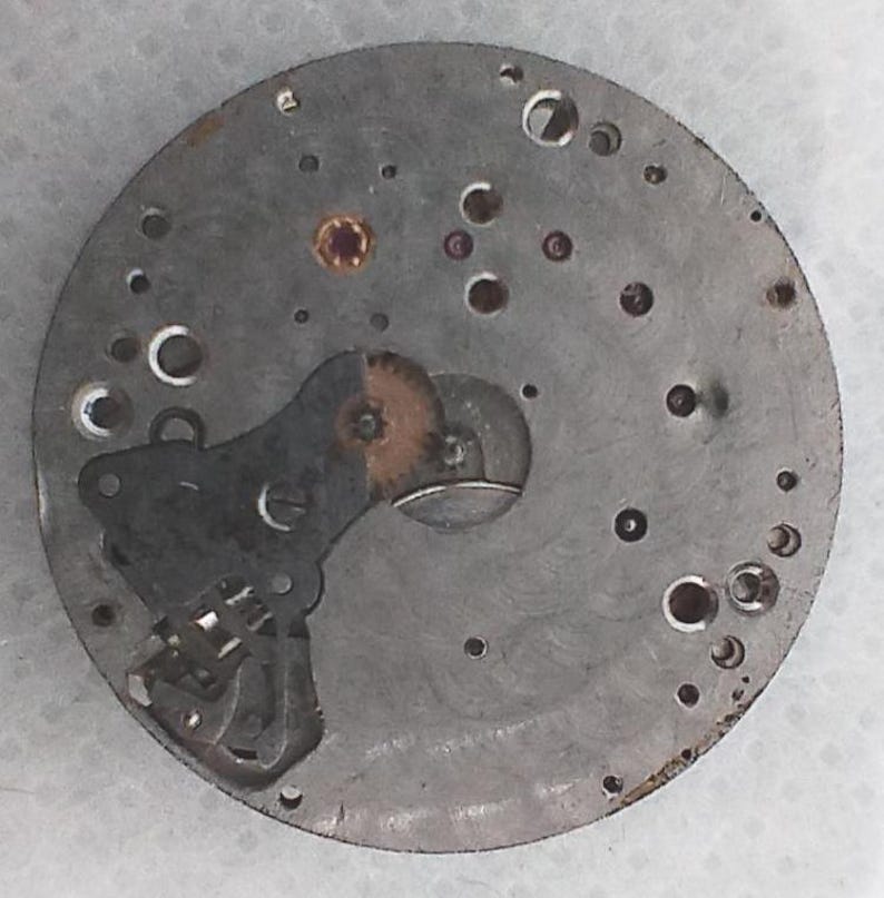 May include: A circular, metallic watch movement component with numerous small holes and embedded jewels. The component features gears, levers, and other mechanical parts. The metal has a worn, aged appearance, suggesting it is vintage or antique.