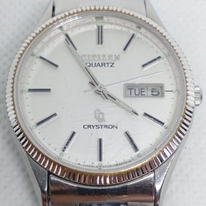 May include: A silver Citizen Quartz Crystron watch with a white face. The watch has a fluted bezel, black hour markers, and a day-date display showing "TUE 5". The watch band is silver.