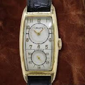 May include: A gold-toned rectangular wristwatch with a white face and black numerals. The watch features two circular dials and a black leather band. The watch case measures approximately 1.2 inches in length.