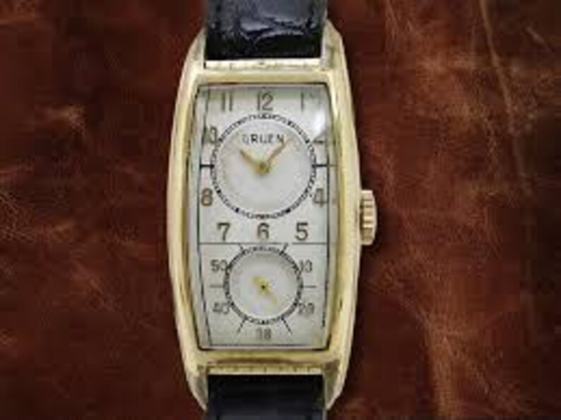 May include: A gold-toned rectangular wristwatch with a white face and black numerals. The watch features two circular dials and a black leather band. The watch case measures approximately 1.2 inches in length.