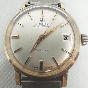 Puede incluir: Reloj vintage Pronto Tropic Master con caja dorada y esfera plateada. El reloj presenta agujas e indicadores dorados, una ventana de fecha a las 3 en punto y el texto "17 Jewels". La correa es de metal.