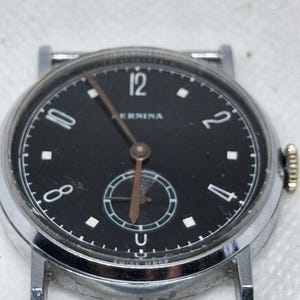 以下が含まれることがあります： 「AERNINA」および「SWISS MADE」の文字が記された黒い文字盤を持つシルバーカラーの腕時計。12時、10時、8時、4時、2時の位置に白いアラビア数字があり、6時の位置に小さなサブダイヤル、細い茶色の針が配置されています。