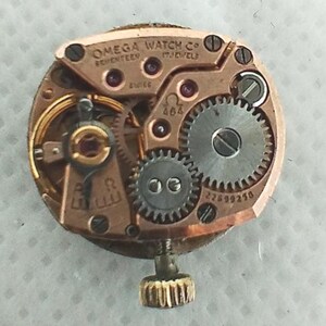 Op de afbeelding: Close-up van een Omega uurwerk. Het mechanisme heeft gouden en zilveren tandwielen, rode juwelen en de tekst "OMEGA WATCH CO" en "SWISS". Het uurwerk heeft een goudkleurige kroon.