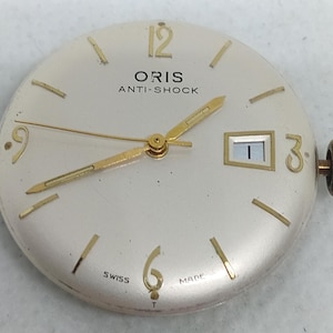 May include: A vintage Oris watch face with a cream dial. Gold hands and hour markers are visible, along with the text "ORIS ANTI-SHOCK" and "SWISS MADE". The date window shows the number 1. The watch has a gold crown.