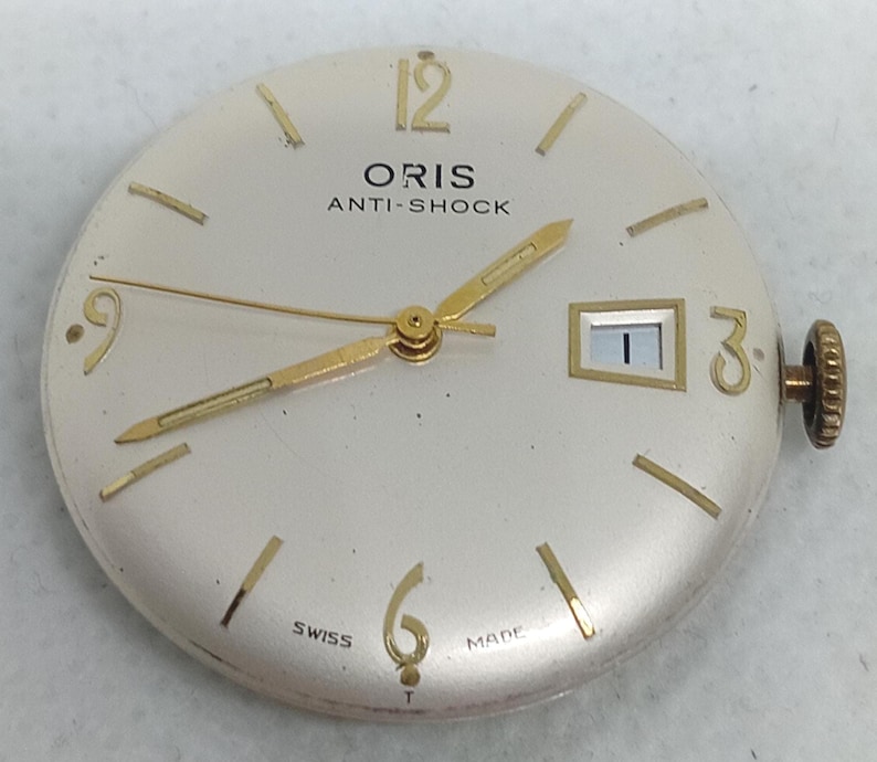 May include: A vintage Oris watch face with a cream dial. Gold hands and hour markers are visible, along with the text "ORIS ANTI-SHOCK" and "SWISS MADE". The date window shows the number 1. The watch has a gold crown.
