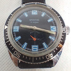 Puede incluir: Un reloj Sporting vintage con esfera negra, marcadores de hora y agujas blancos y azules. El reloj tiene un bisel negro con marcas blancas y una correa de cuero negro. El texto "SPORTING 15 RUBIS ANTICHOC" es visible en la esfera.